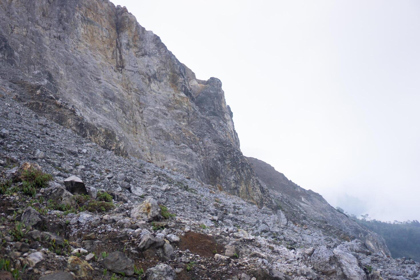 A rugged rocky mountainside under a cloudy sky, showing steep, barren cliffs with loose stones scattered across the terrain. Minimal vegetation clings to the inhospitable environment. photo