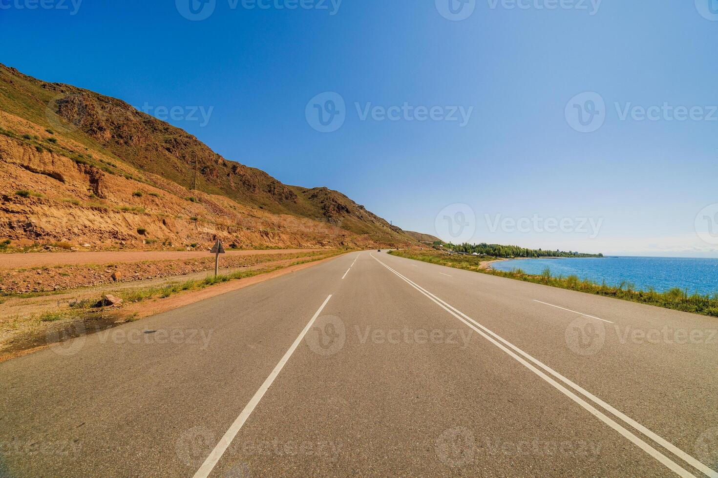 asphalt interstate road between yellow sedimentary rock mountain and large lake at sunny summer day photo