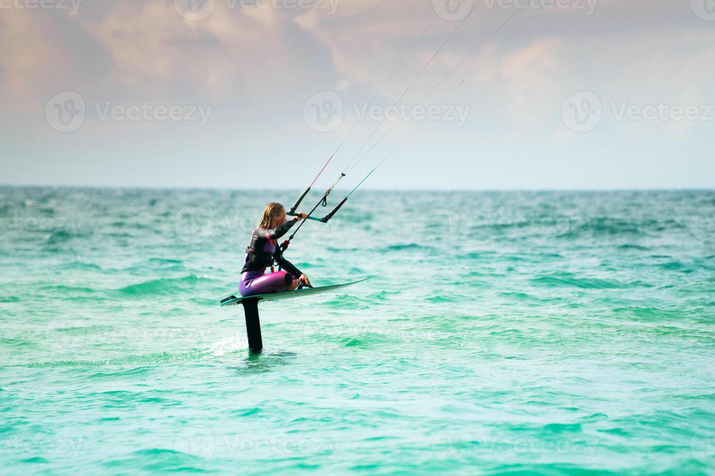 A young athlete in a colorful wetsuit sits on her hydrofoil and rides the waves with kitesurfing. Shot with a telephoto lens photo