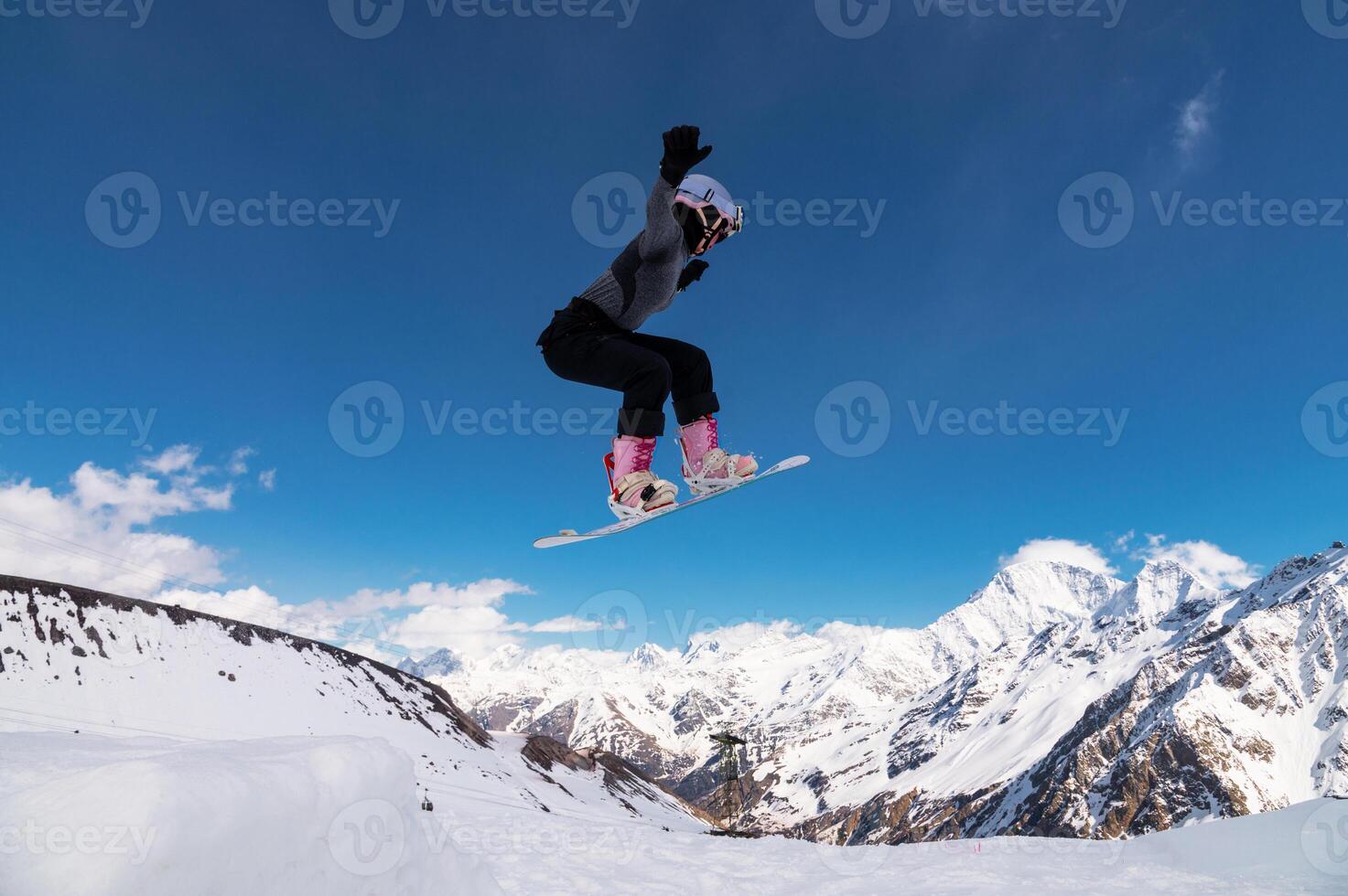 Female snowboarder jumping off a ski jump and doing a trick. Amazing shot of a snowboarder flying in the air and grabbing a trick against the backdrop of snowy mountains photo