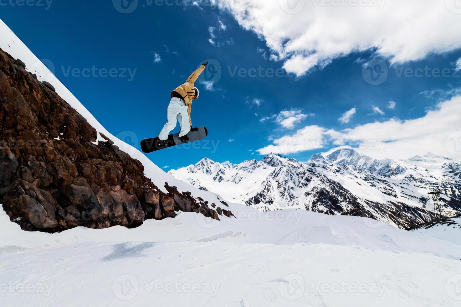 Professional snowboarder in helmet and ski clothes doing a trick in a jump against the background of high snowy mountains. Wide angle. Sports shooting photo