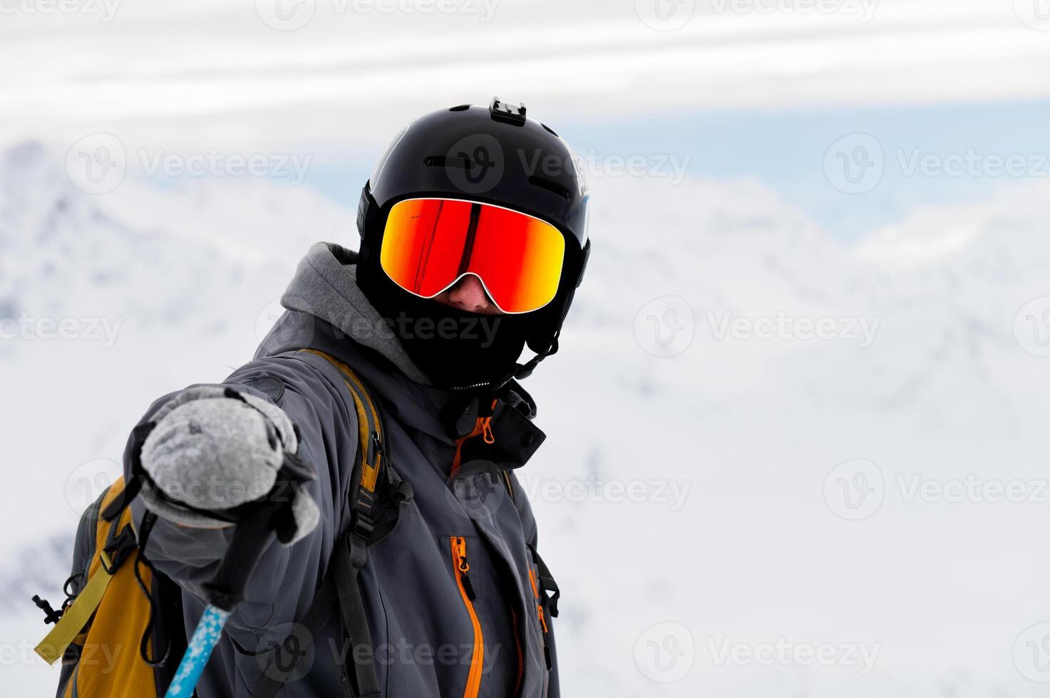 man points his hand at the camera in sunglasses with a ski helmet on his head and holds a stick in his hand against the background of white snow and mountains photo