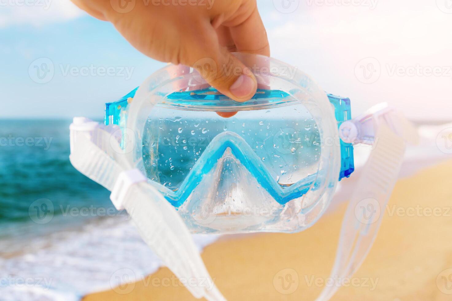 A man holds a snorkel mask in his hand against a clear blue sky and turquoise ocean. Diving mask in a man's hand. Photo of a mask and snorkel for swimming in a pool or sea with a central composition