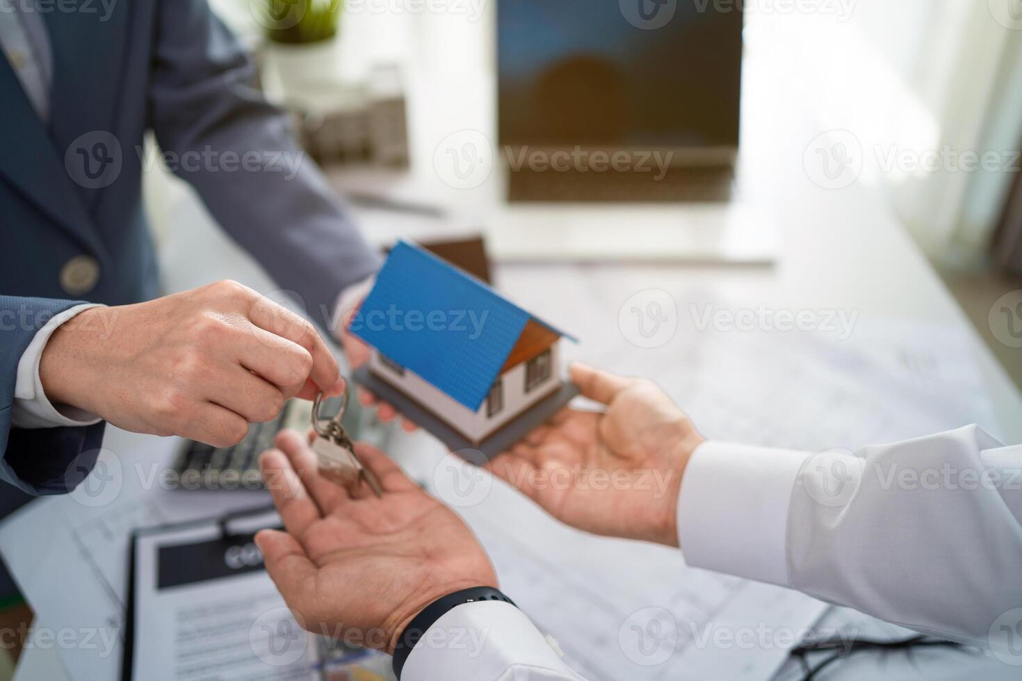 A man is handing a key to a woman. The woman is holding a blue house model. The man is wearing a suit and tie photo