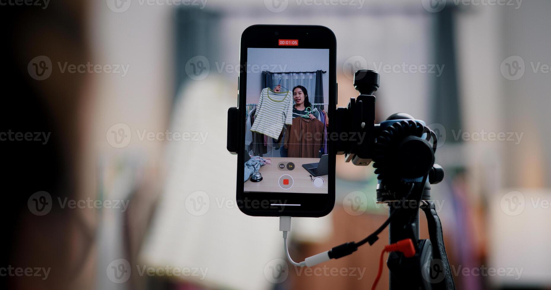Selective focus on mobile phone of Cheerful woman influencer is live streaming sell clothes on social media or blogging in home studio. Remote buying, Online shopping. photo