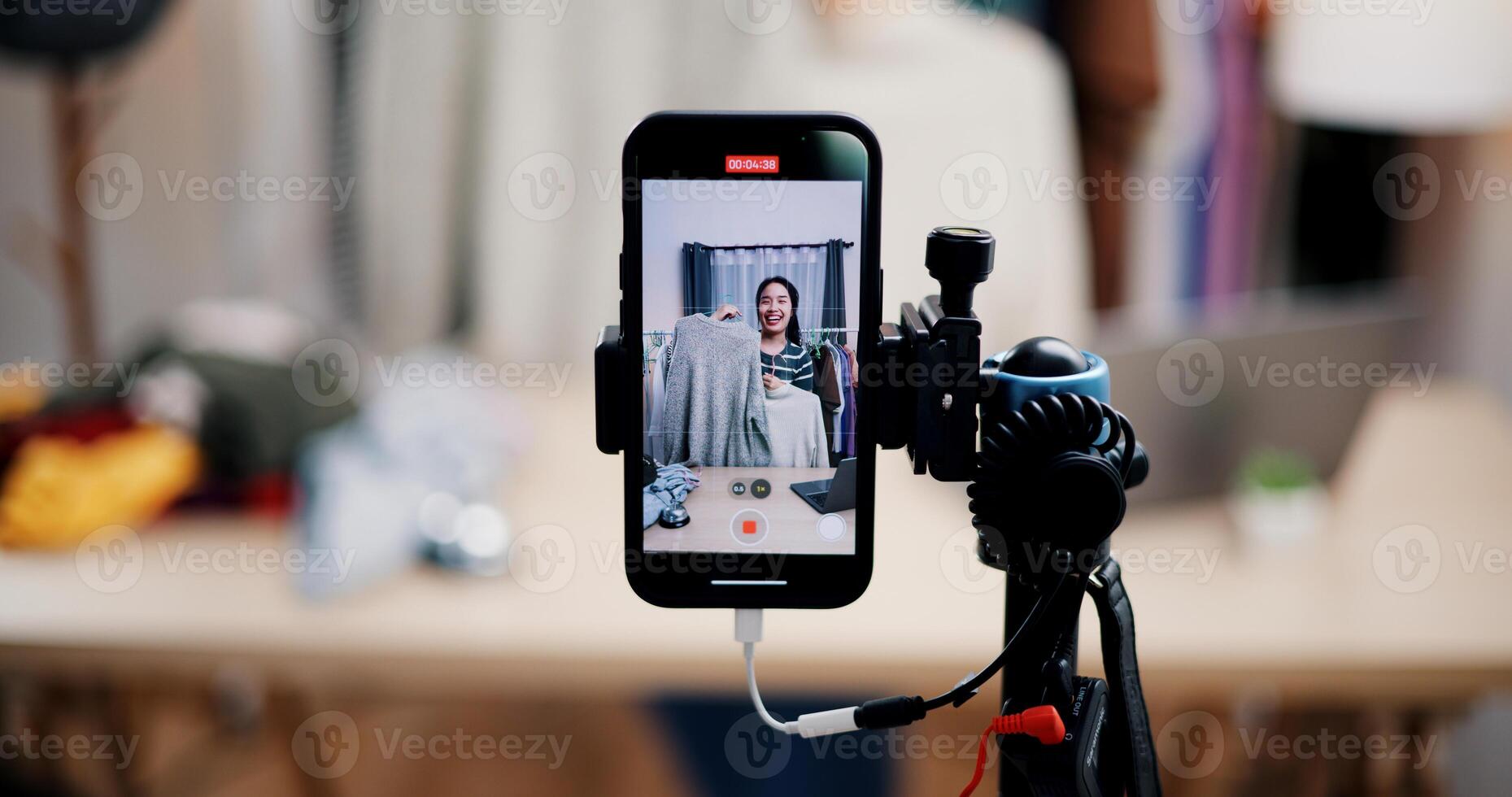 Selective focus on mobile phone of Cheerful woman influencer is live streaming sell clothes on social media or blogging in home studio. Remote buying, Online shopping. photo