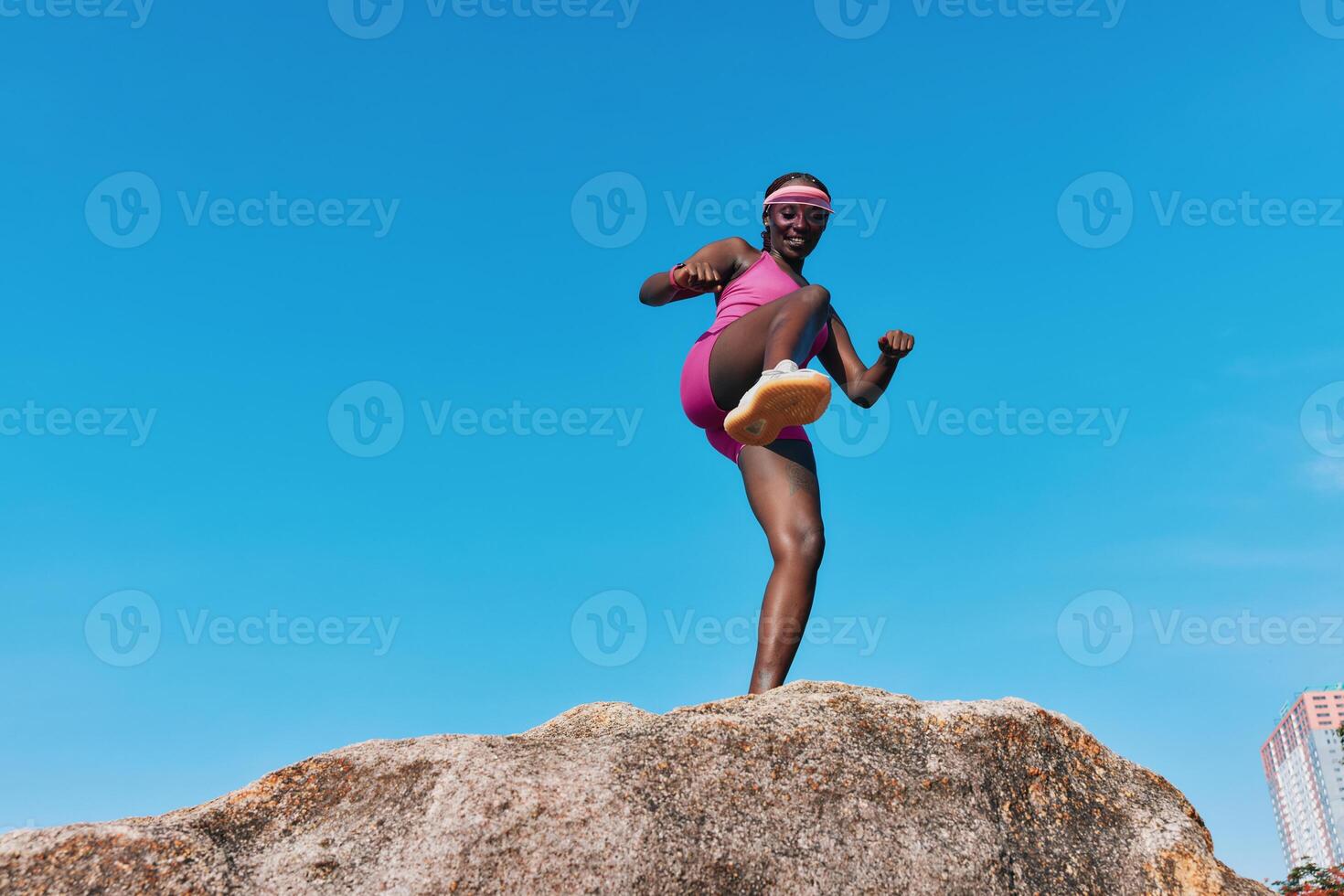 Energetic female athlete poses on a large rock beneath a vibrant blue sky Her expression exudes photo