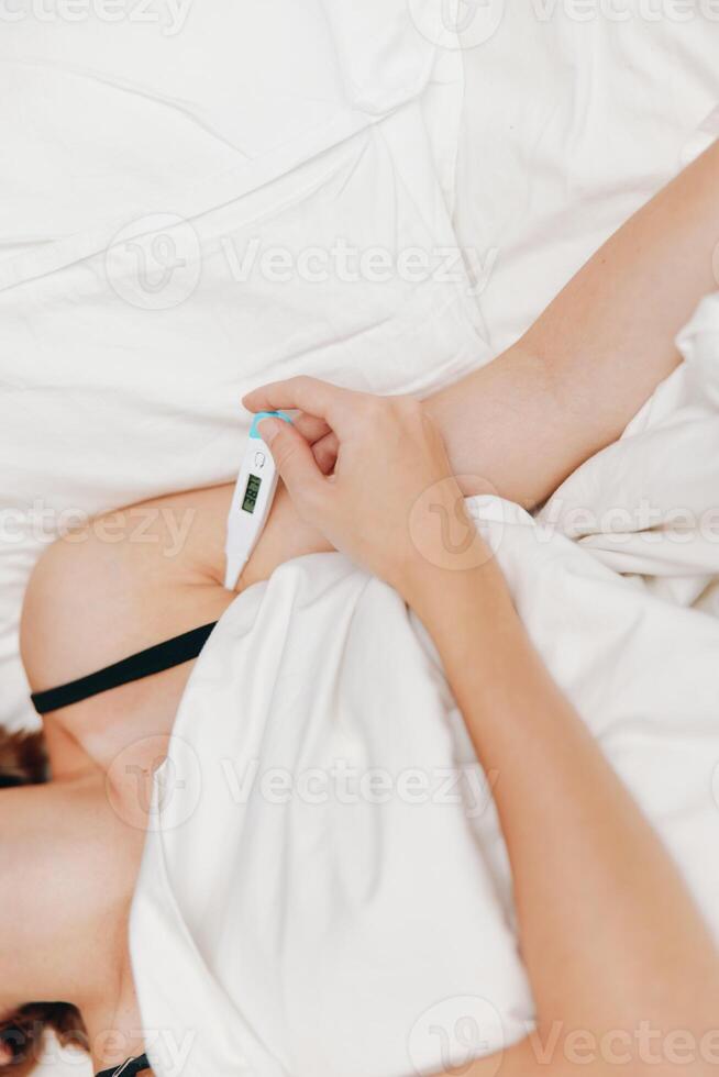 Ill woman lying in bed holding a thermometer, with another thermometer placed next to her head. a photo
