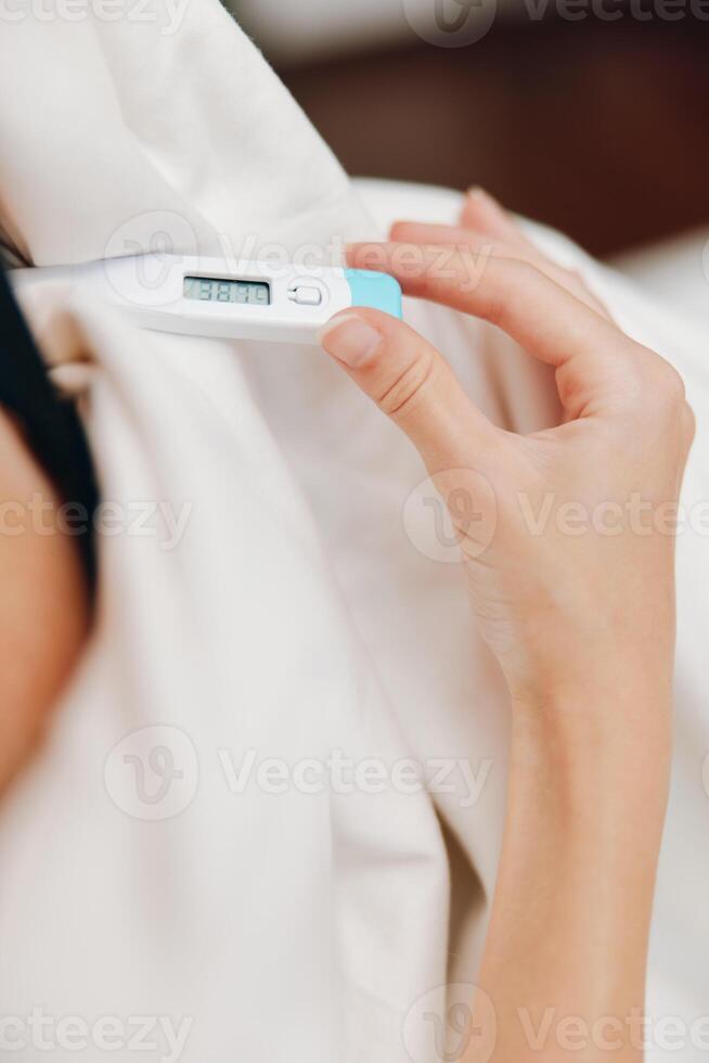 Female using digital thermometer while resting in bed covered with blanket. photo