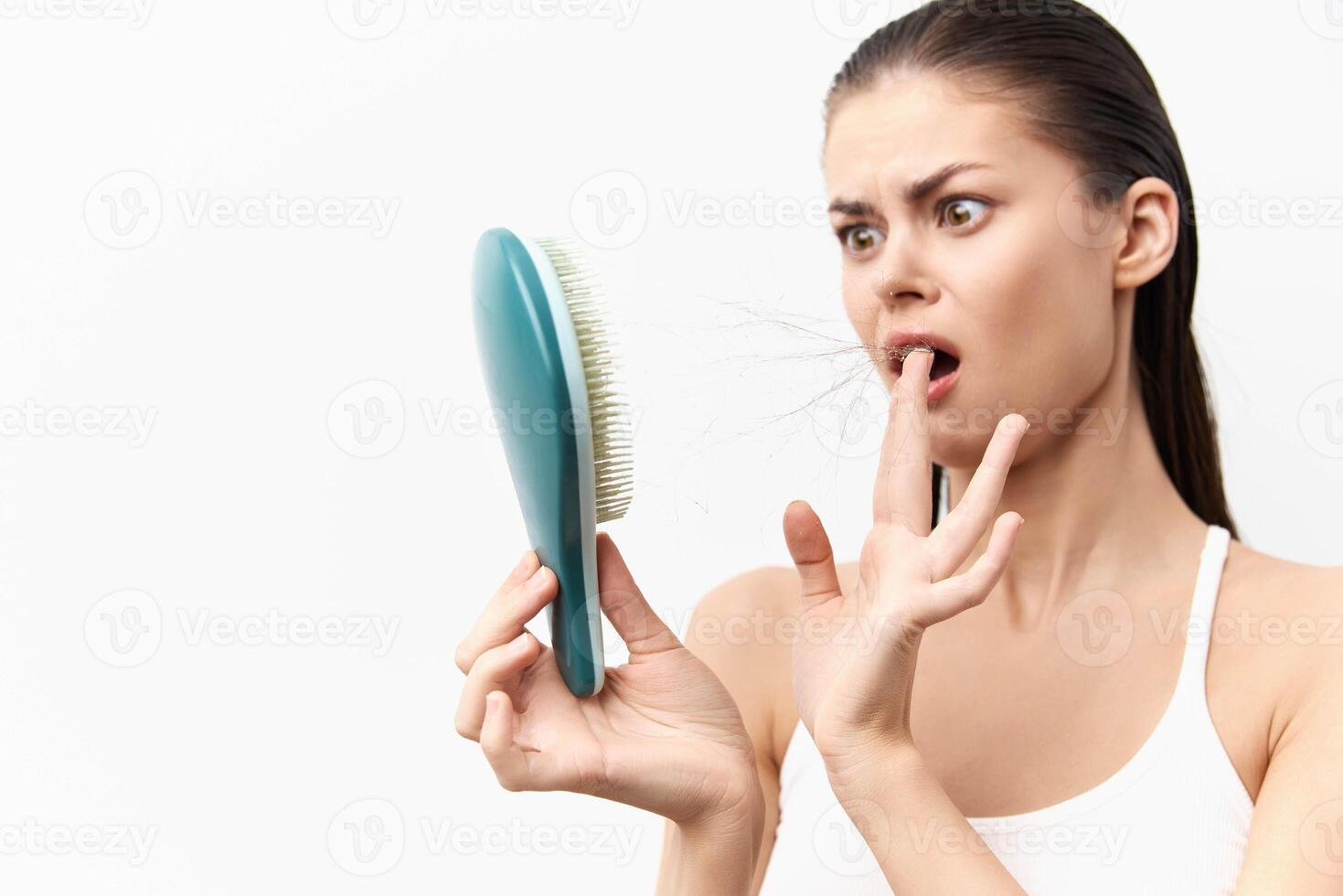 A surprised young woman is gazing at a hairbrush filled with hair in her hand against a simple white photo