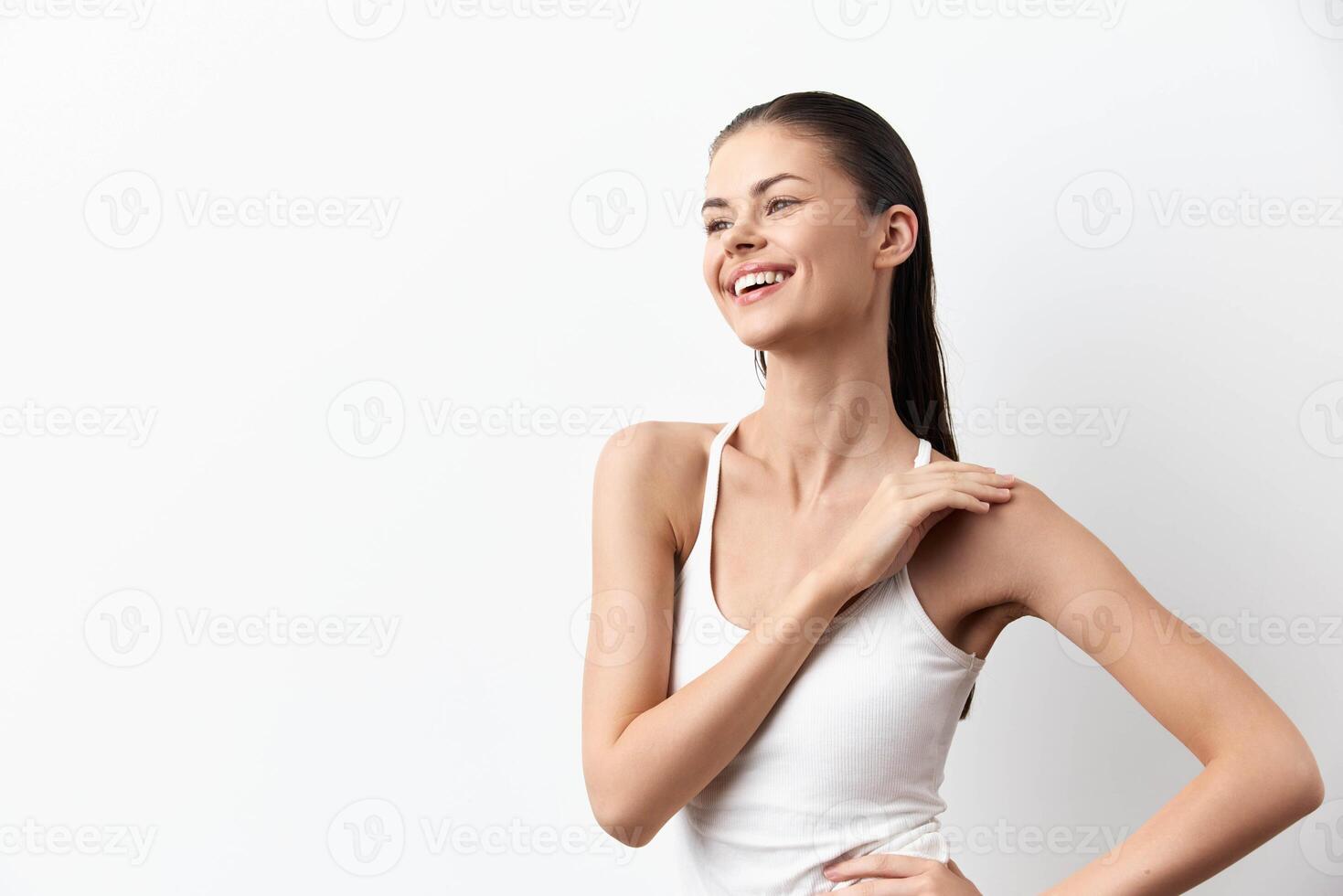 A cheerful young woman in a white tank top smiles warmly against a bright background, radiating photo