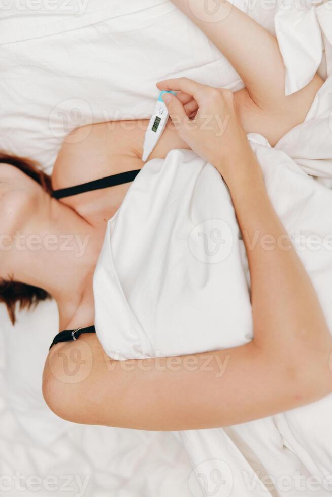 Ill woman lying in bed with thermometer, covered by white sheet and pillow. photo