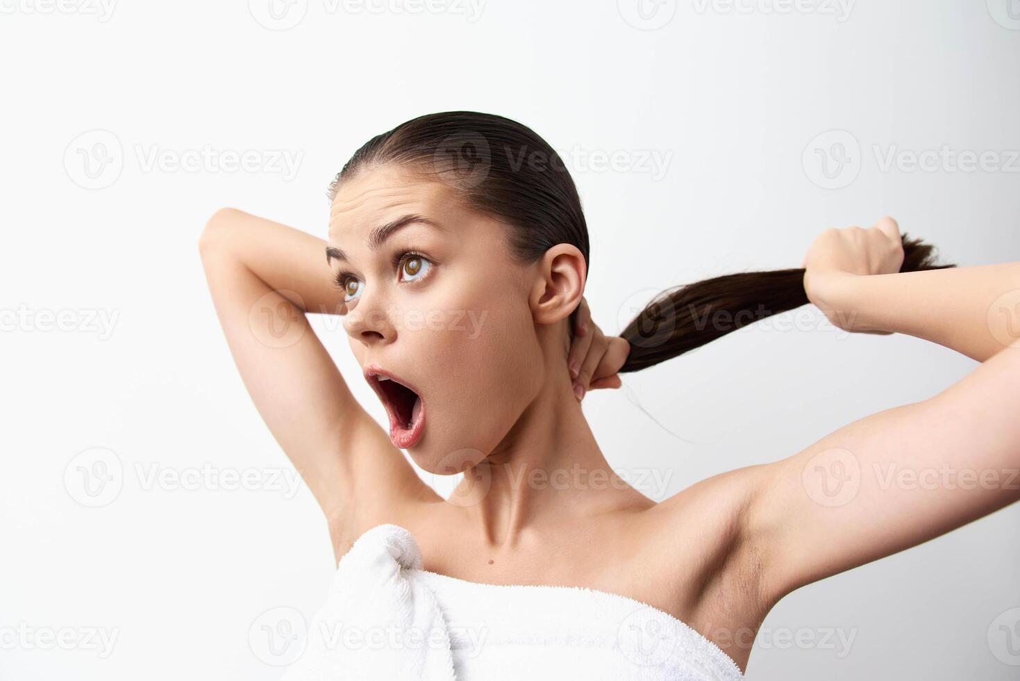 A young woman wrapped in a towel looks surprised while pulling her hair back, set against a plain photo