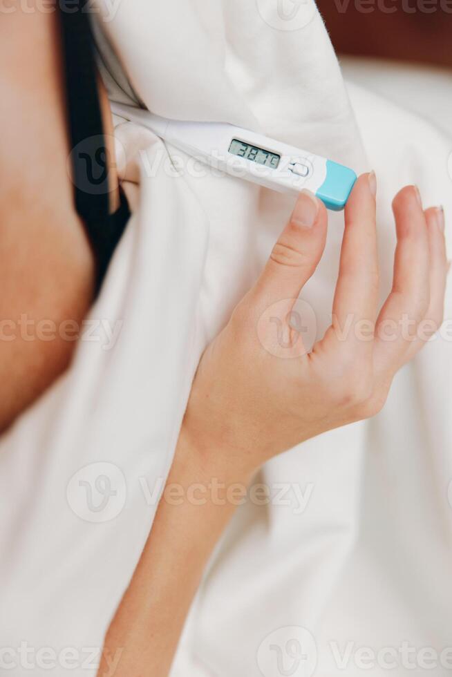 Woman holding electronic thermometer in bed under white sheet and blanket. checking temperature in a photo