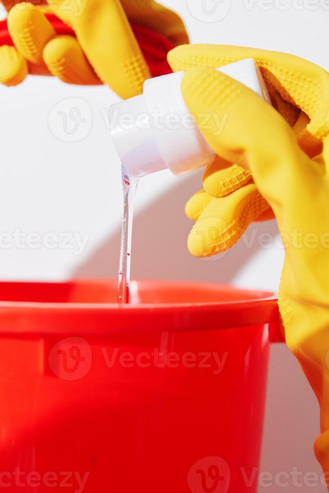 Yellow-gloved woman pouring liquid into red bucket next to white container. photo