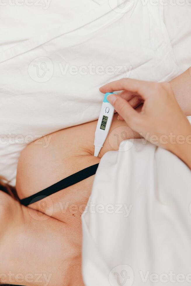 Woman laying in bed, holding an electronic thermometer while another thermometer is placed next to photo