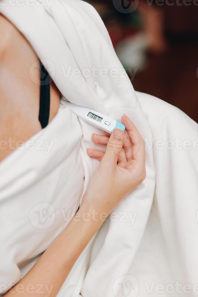Ill woman lying in bed with a thermometer, covered with a blanket and pillow. photo
