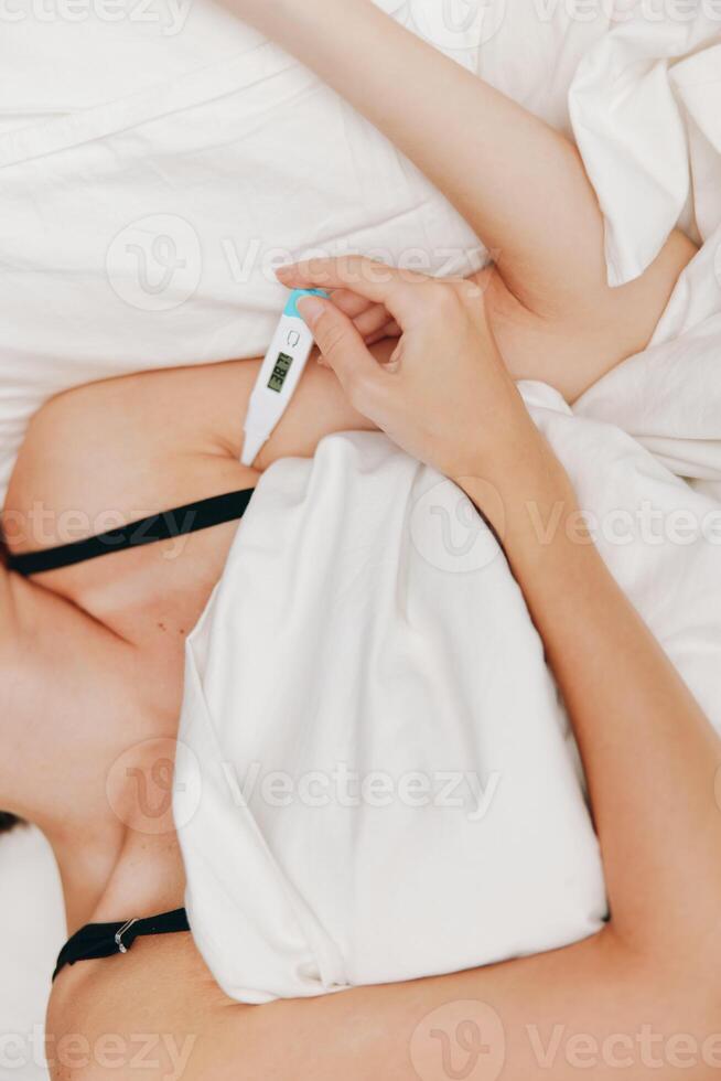 Ill woman lying in bed with a thermometer in her mouth, another one lying on the bedside table next photo
