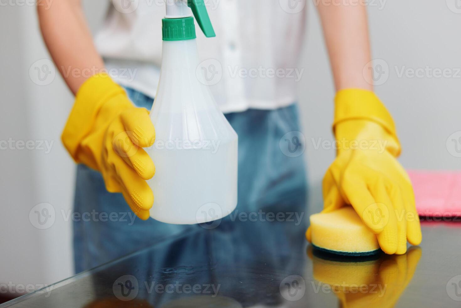 Woman in yellow rubber gloves holding cleaning solution and sponge on glass table in front of photo