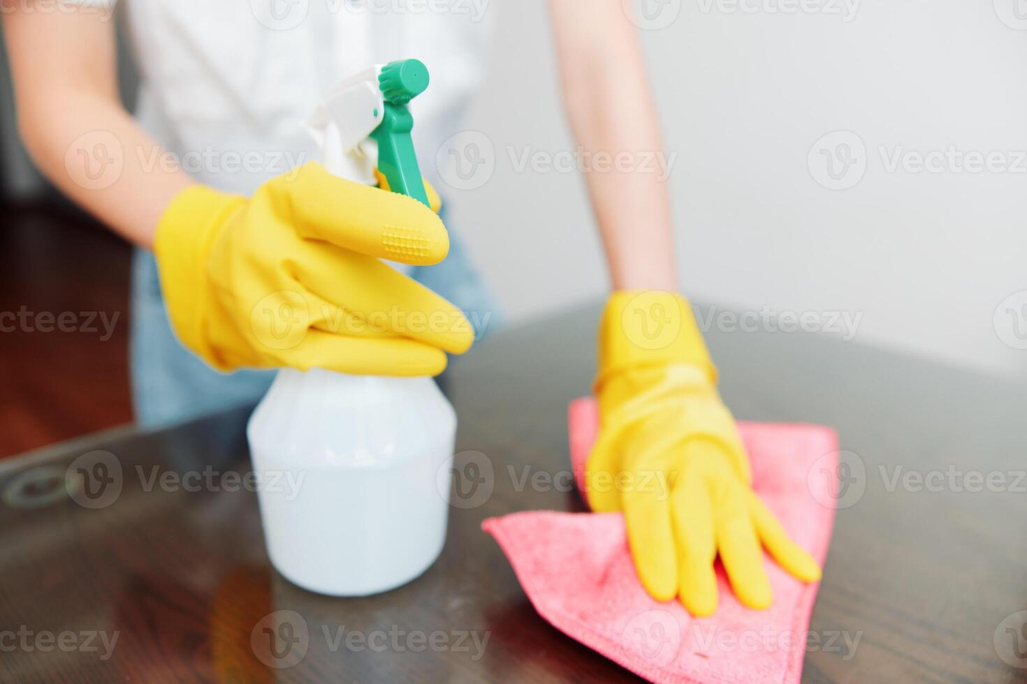 Woman cleaning table with yellow gloves using bottle of solution and rag on wooden surface. photo