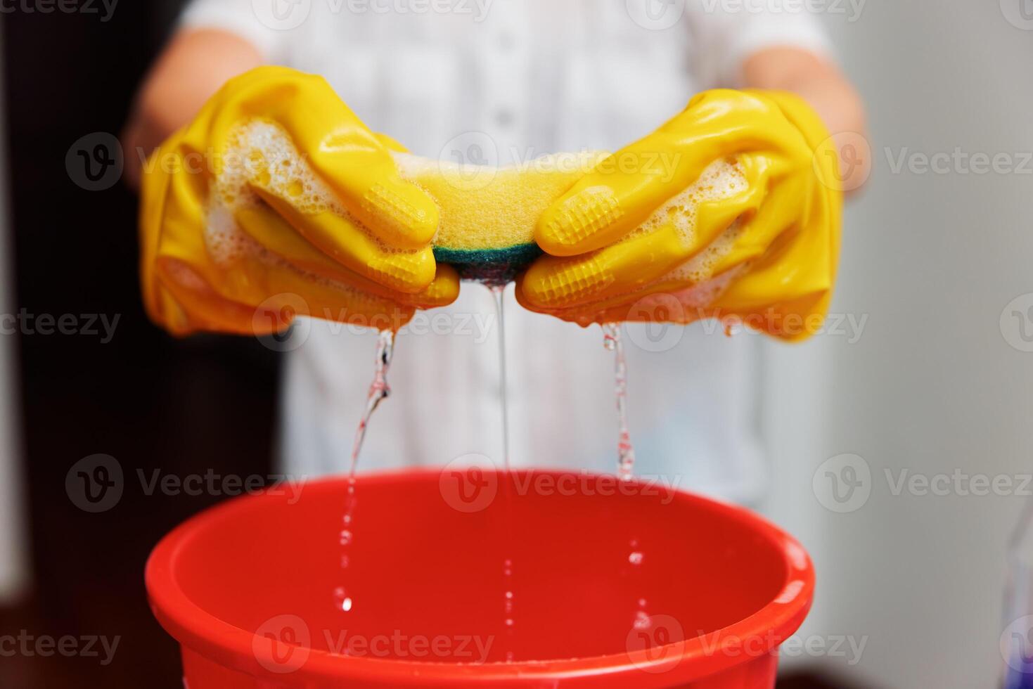 Woman in yellow gloves holding sponge over red bucket with yellow sponge inside. photo