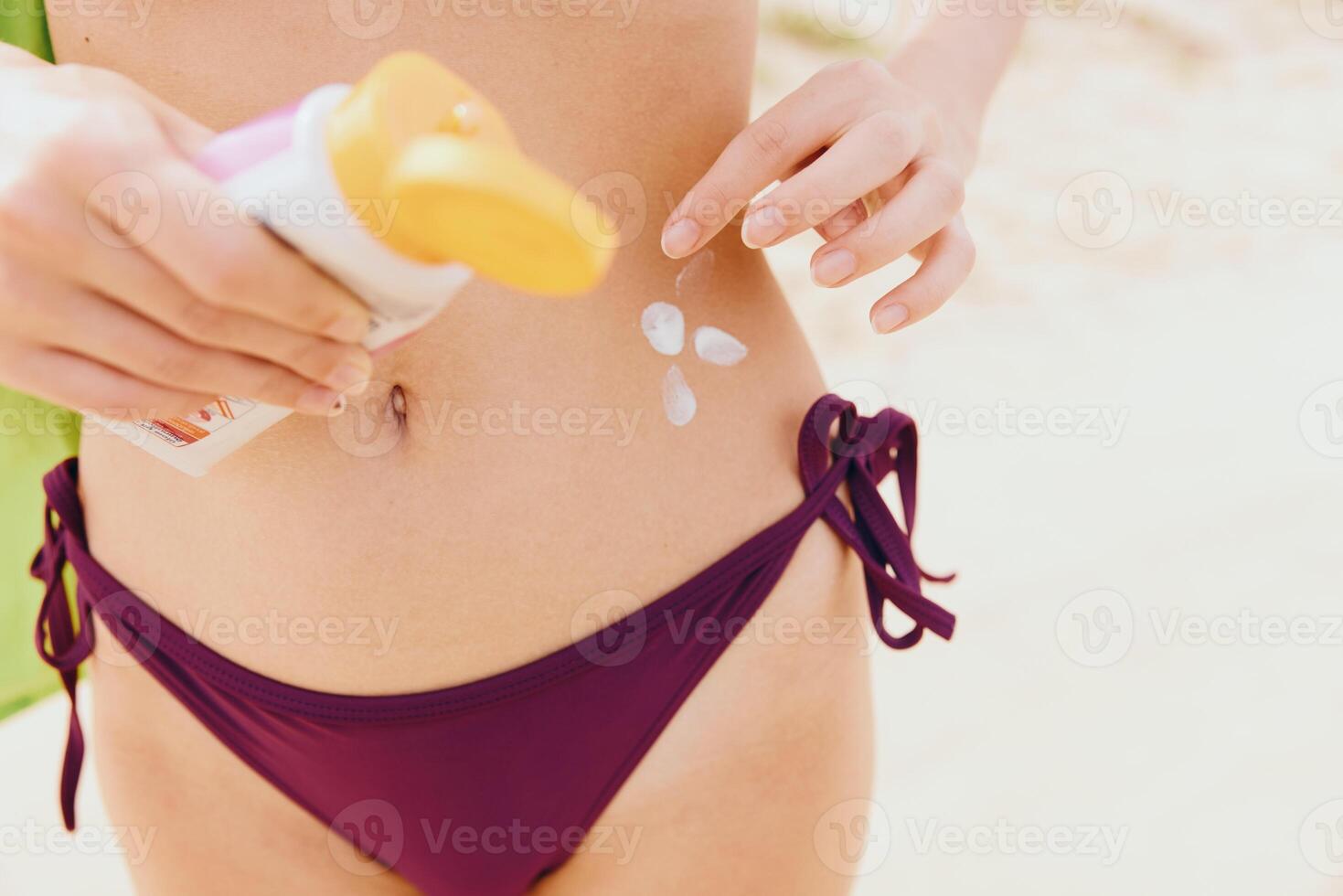 Woman in purple bikini holding sunscreen bottle on stomach, towel covering face, standing on sandy photo