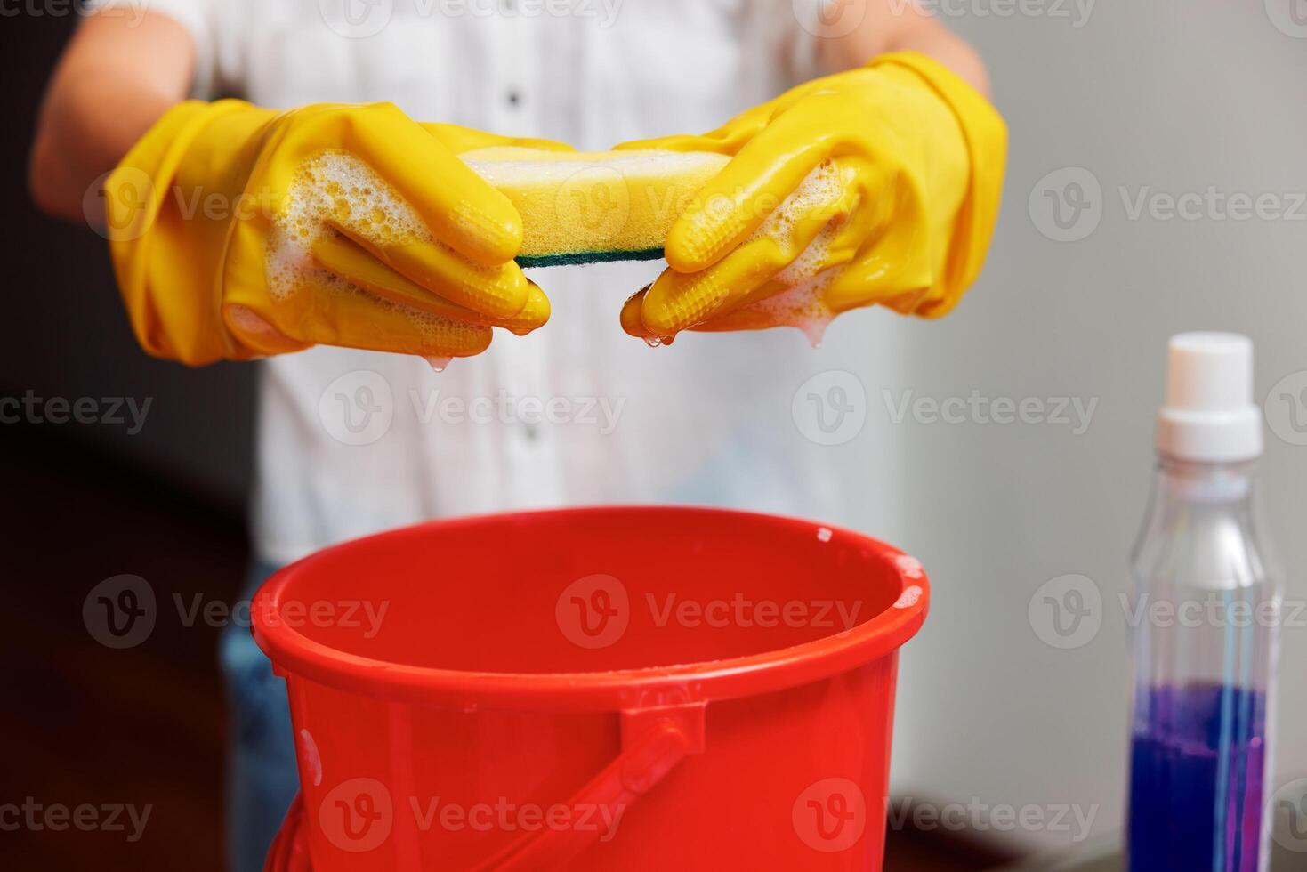 Woman with yellow rubber gloves holding a sponge over a red bucket with a bottle of cleaning photo