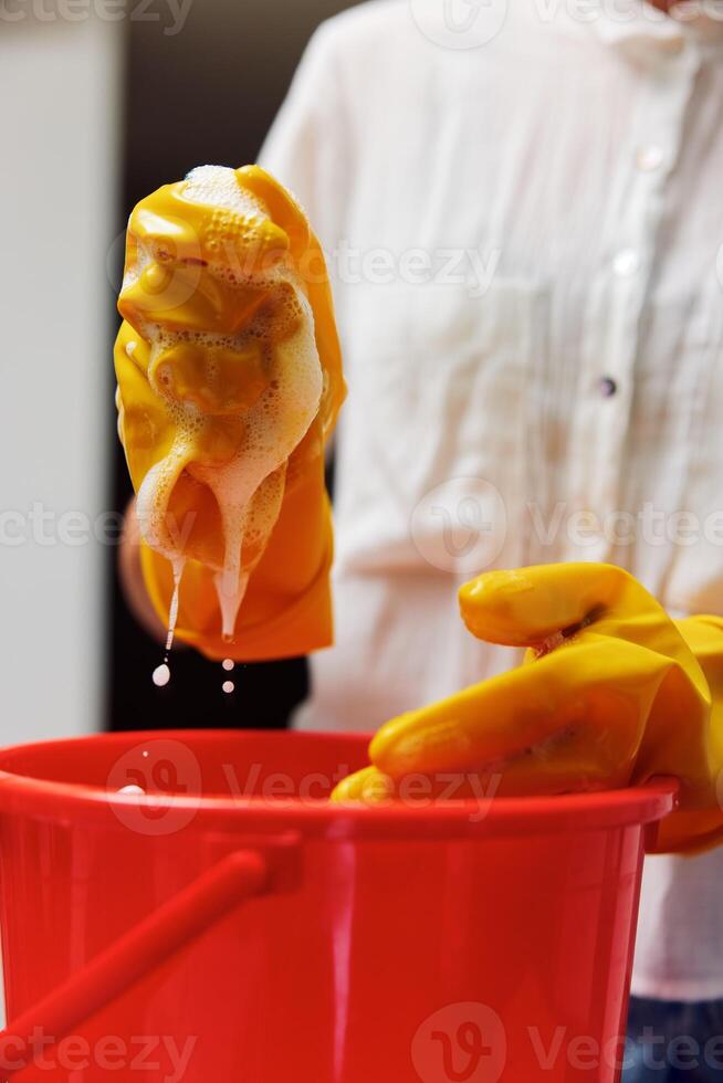Woman in white shirt and yellow rubber gloves cleaning a bucket of soapy water with a sponge. photo