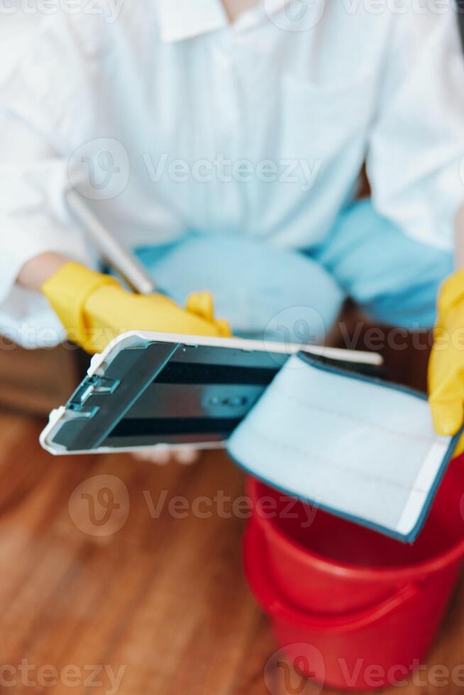Cleaner in white shirt and yellow gloves uses tablet while mopping floor with bucket of water. photo