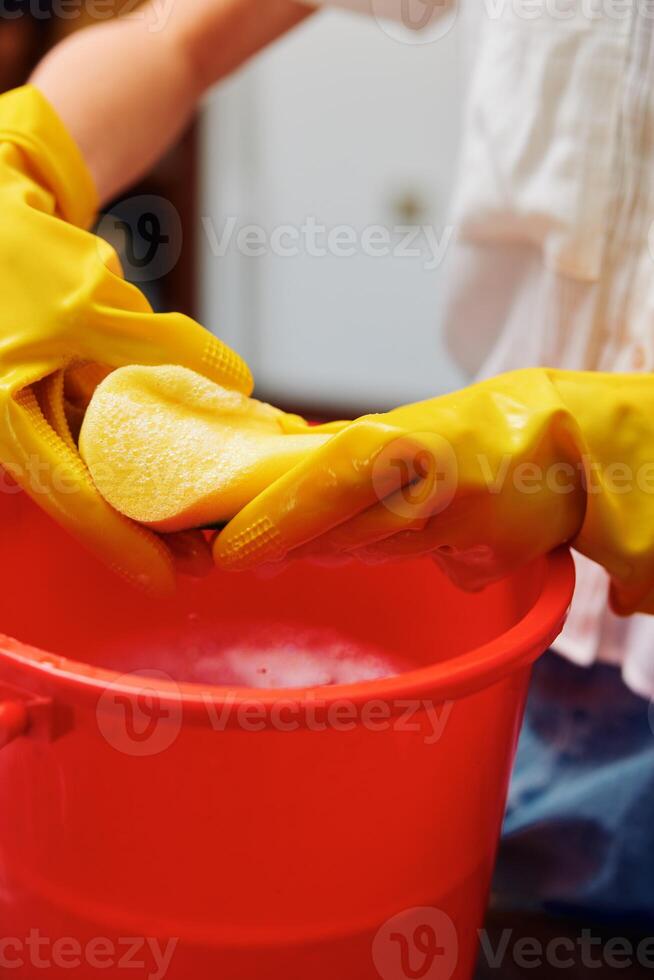 Woman wearing yellow gloves washing an object in a red bucket with a sponge in the sink. photo