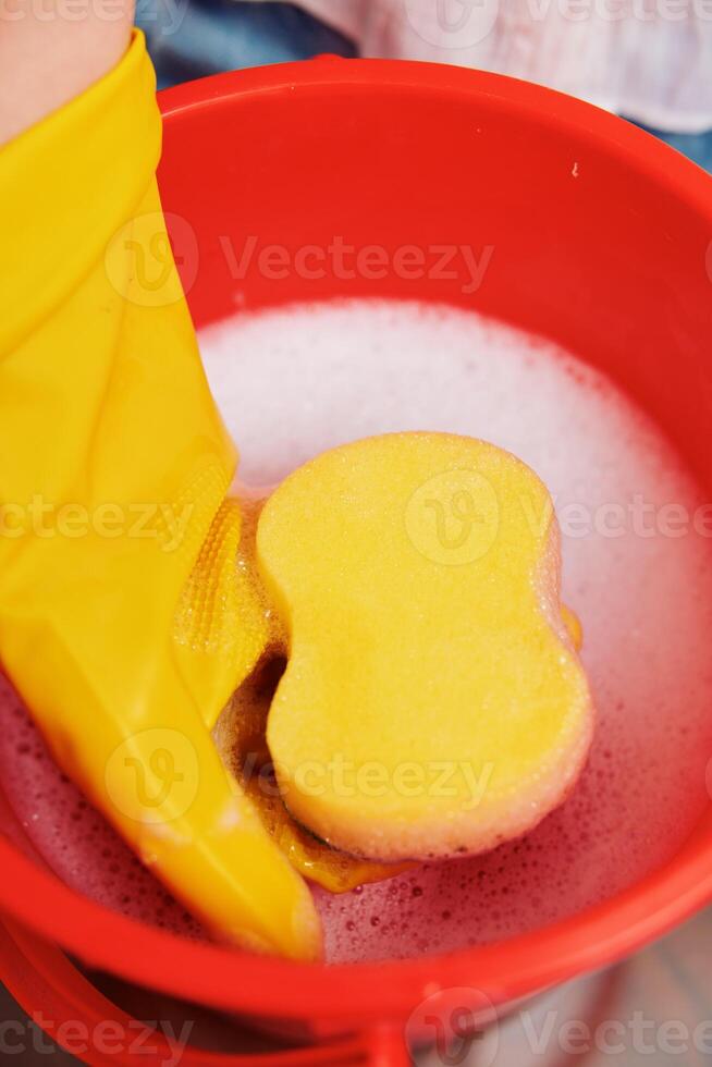 Cleaning woman in yellow glove washing sponge in red bucket in sink. photo