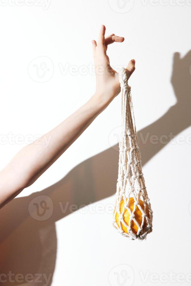 Man holding up a white bag with an orange fruit inside, casting a shadow against a white wall. photo