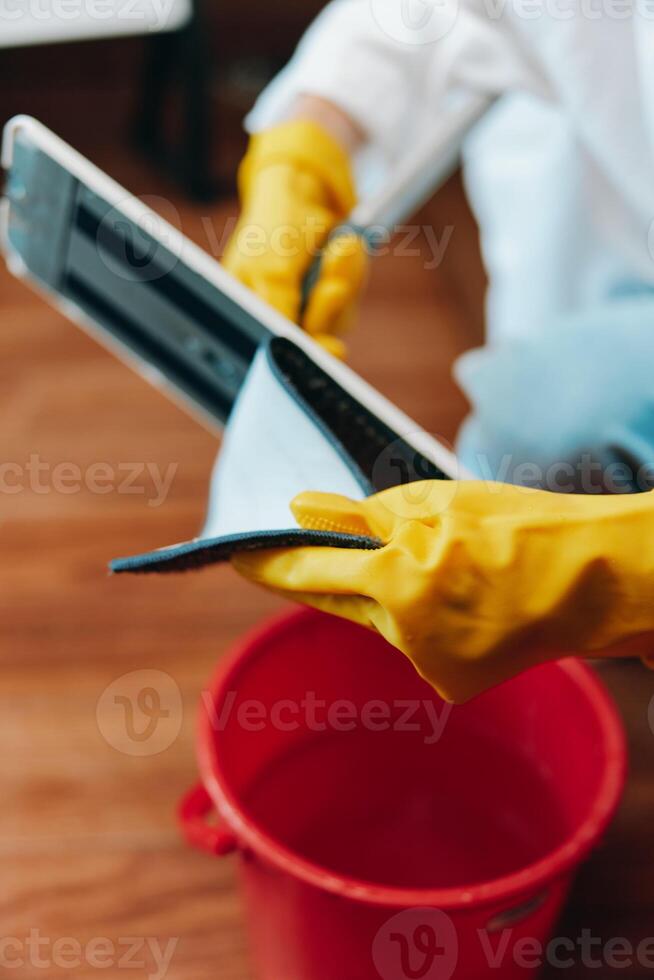 Person wearing yellow rubber gloves using a tablet on a wooden floor next to a bucket of water and photo