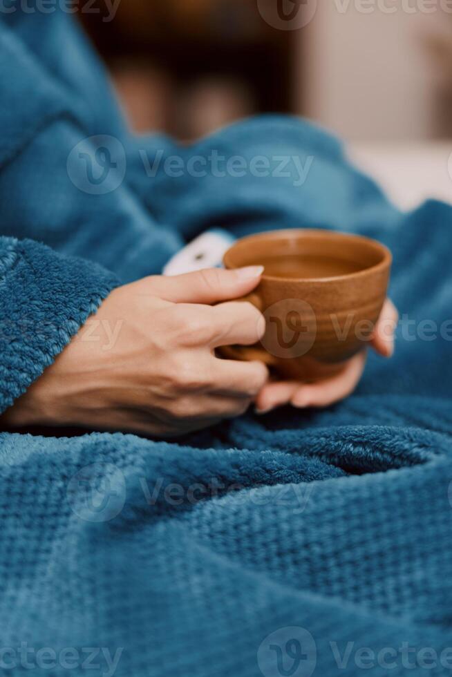 A person wrapped in a blue blanket is holding a cup of coffee in one hand and a remote control in photo
