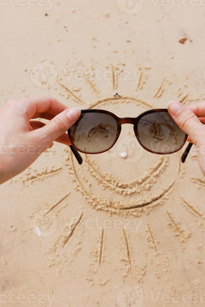 Hands holding sunglasses on a sandy beach with a drawn sun and smiley face. beach accessories photo