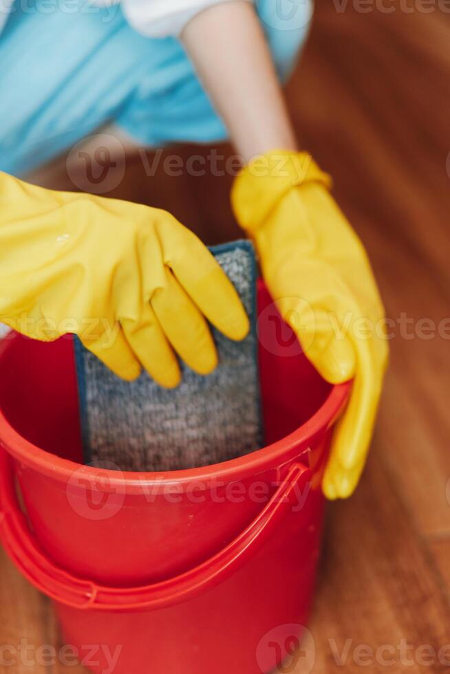 Person in rubber gloves holding bucket with sponge on hardwood floor in front of another person in photo