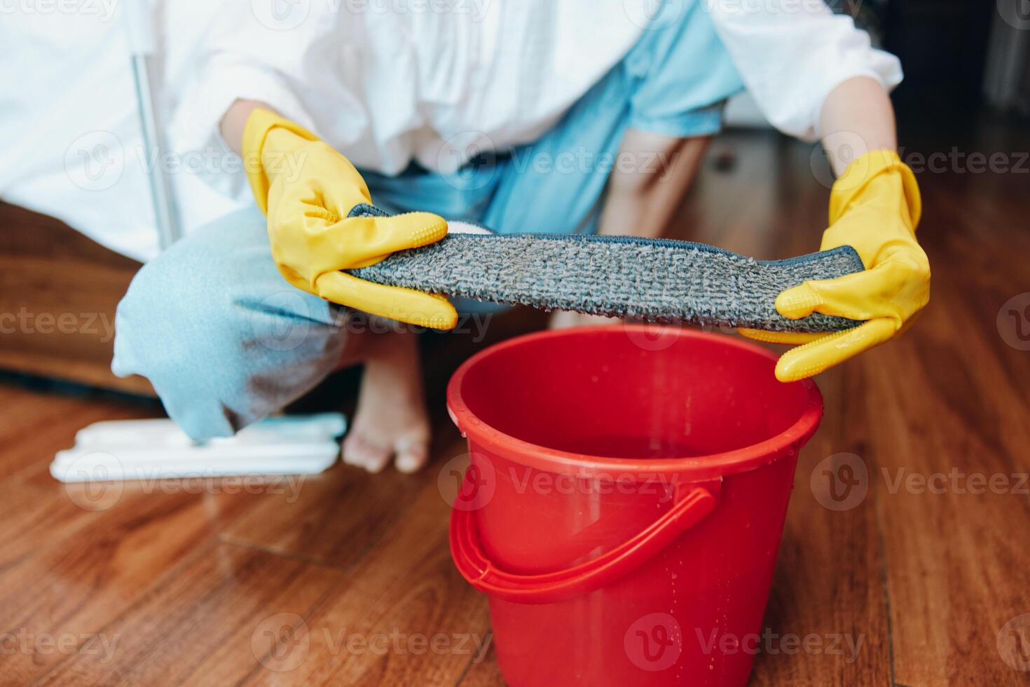 Person wearing rubber gloves using mop and bucket to clean floor in front of red pail. photo