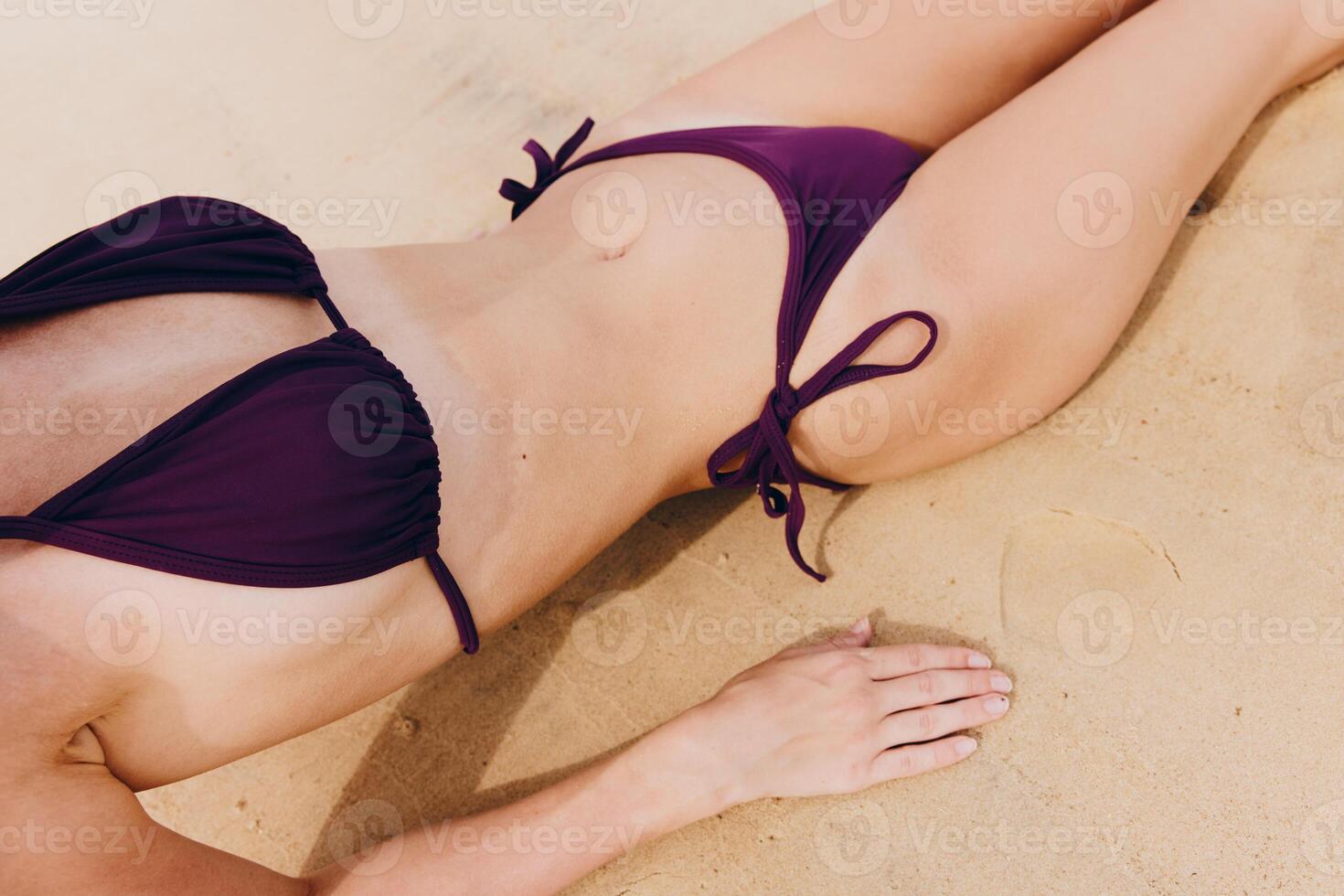 Woman in purple bikini lying on sandy beach with hands on hips and feet crossed in front. photo