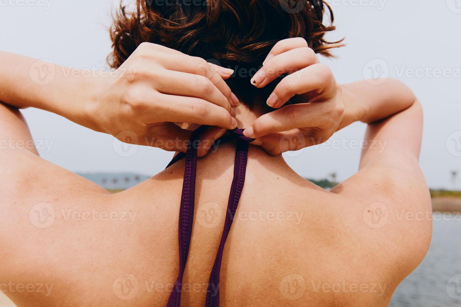 Woman tying a purple tie around her neck near the water, with a body of water in the background. photo