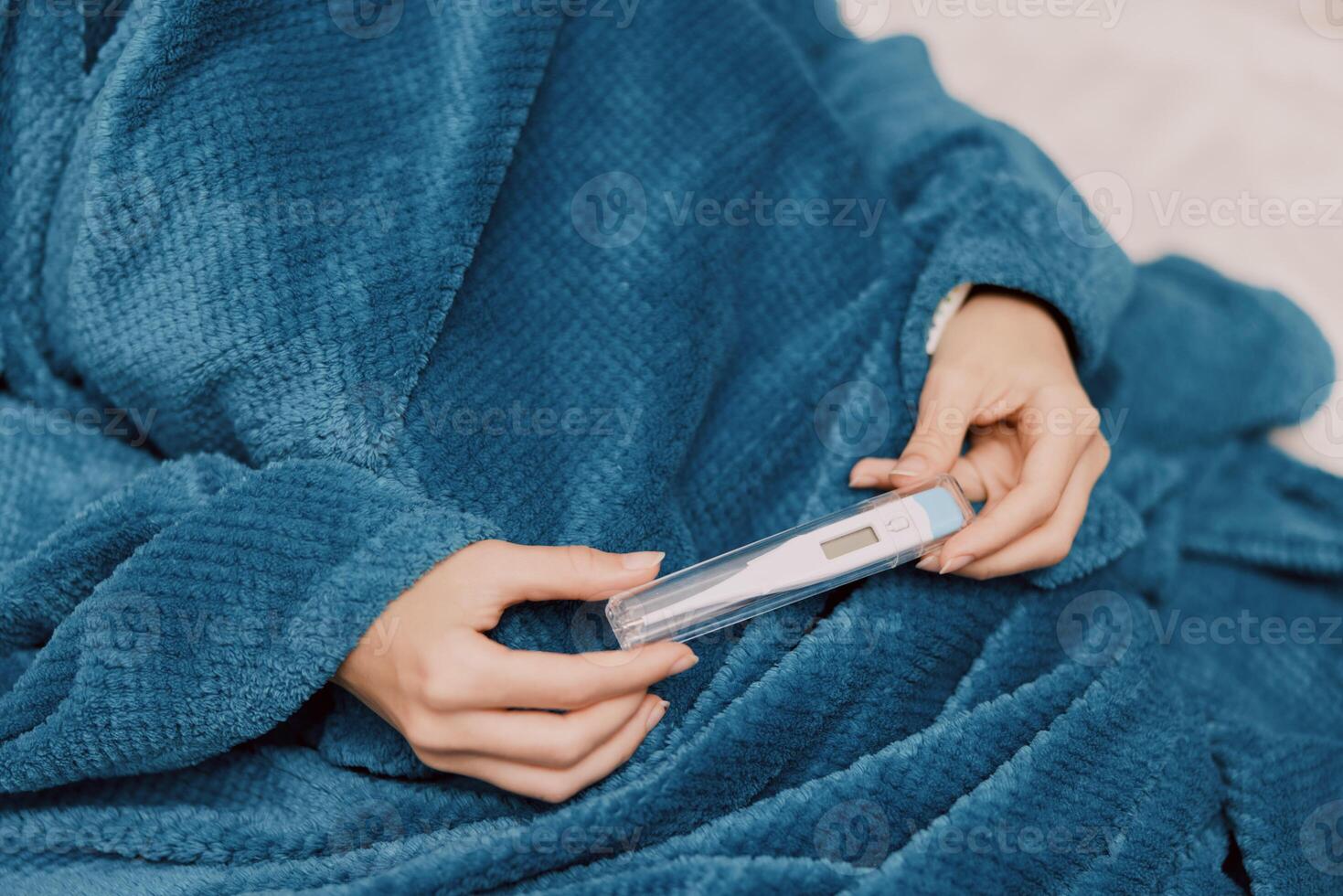 Sick woman sitting on a bed wrapped in a blue blanket, holding a thermometer in her hand. her feet photo
