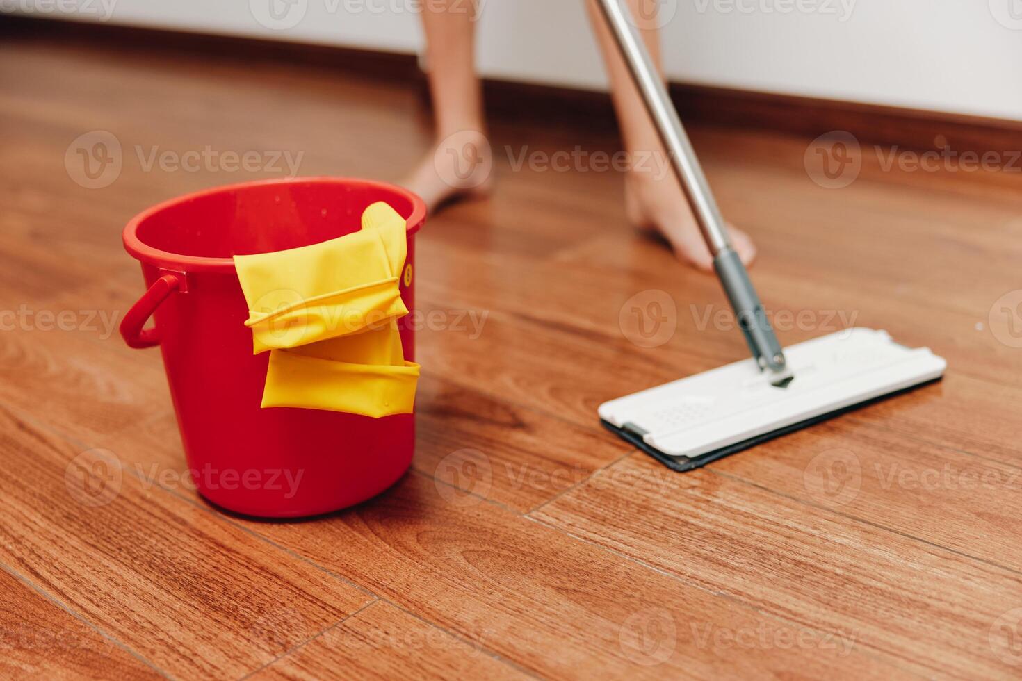 Woman mopping wooden floor in front of red bucket with cleaning supplies. a household chore being photo