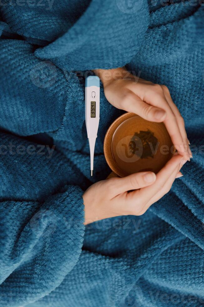 Person wrapped in blanket holding thermometer with cup of tea a person is sitting wrapped in a cozy photo