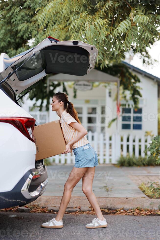 a young woman is lifting a cardboard box into the trunk of a car, surrounded by greenery on a sunny photo
