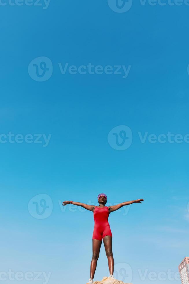 A confident woman stands with arms outstretched on a rock, wearing a red swimsuit, with an expansive photo