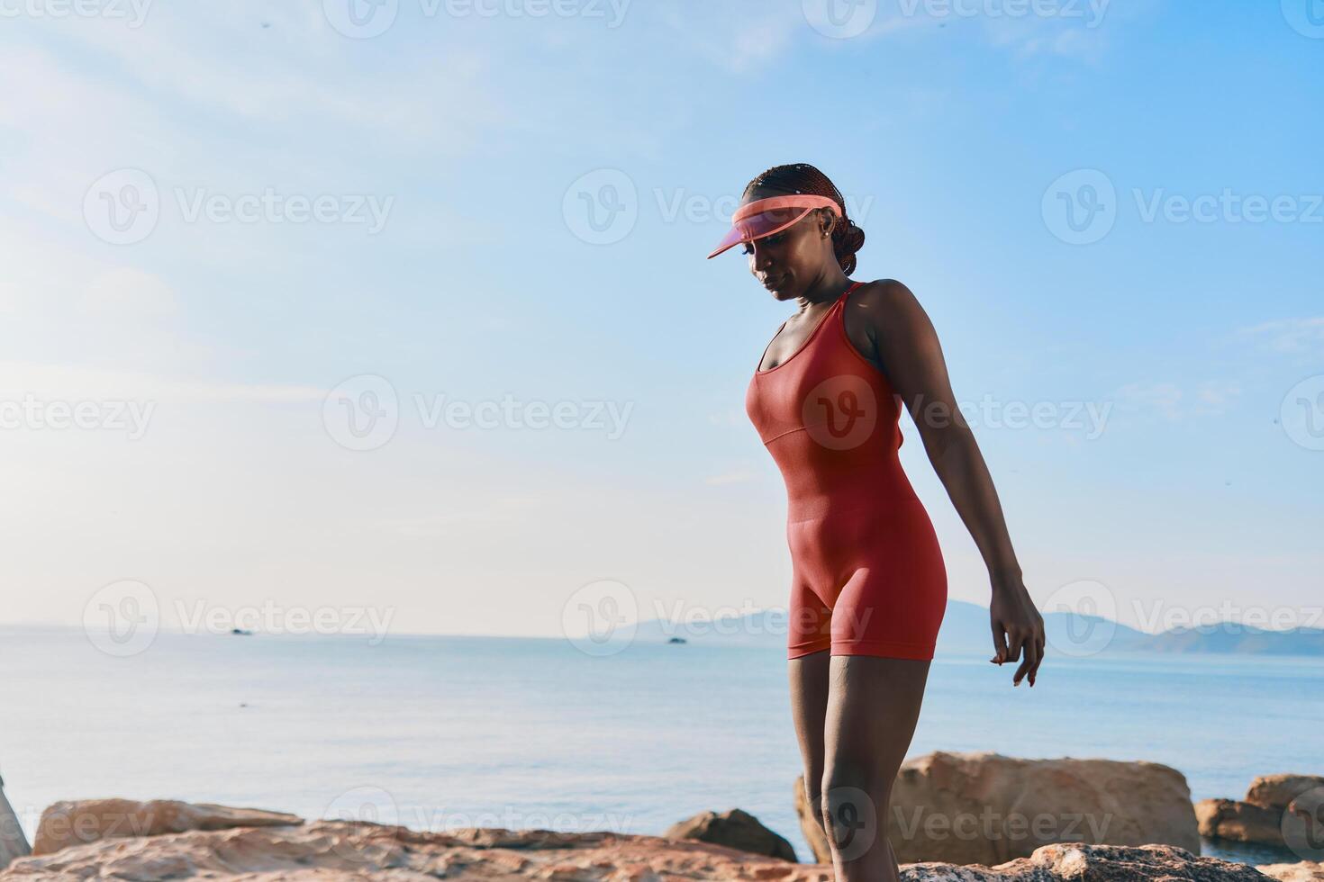 A confident woman in a red swimsuit, wearing a visor, enjoys a peaceful stroll along the beach, with photo
