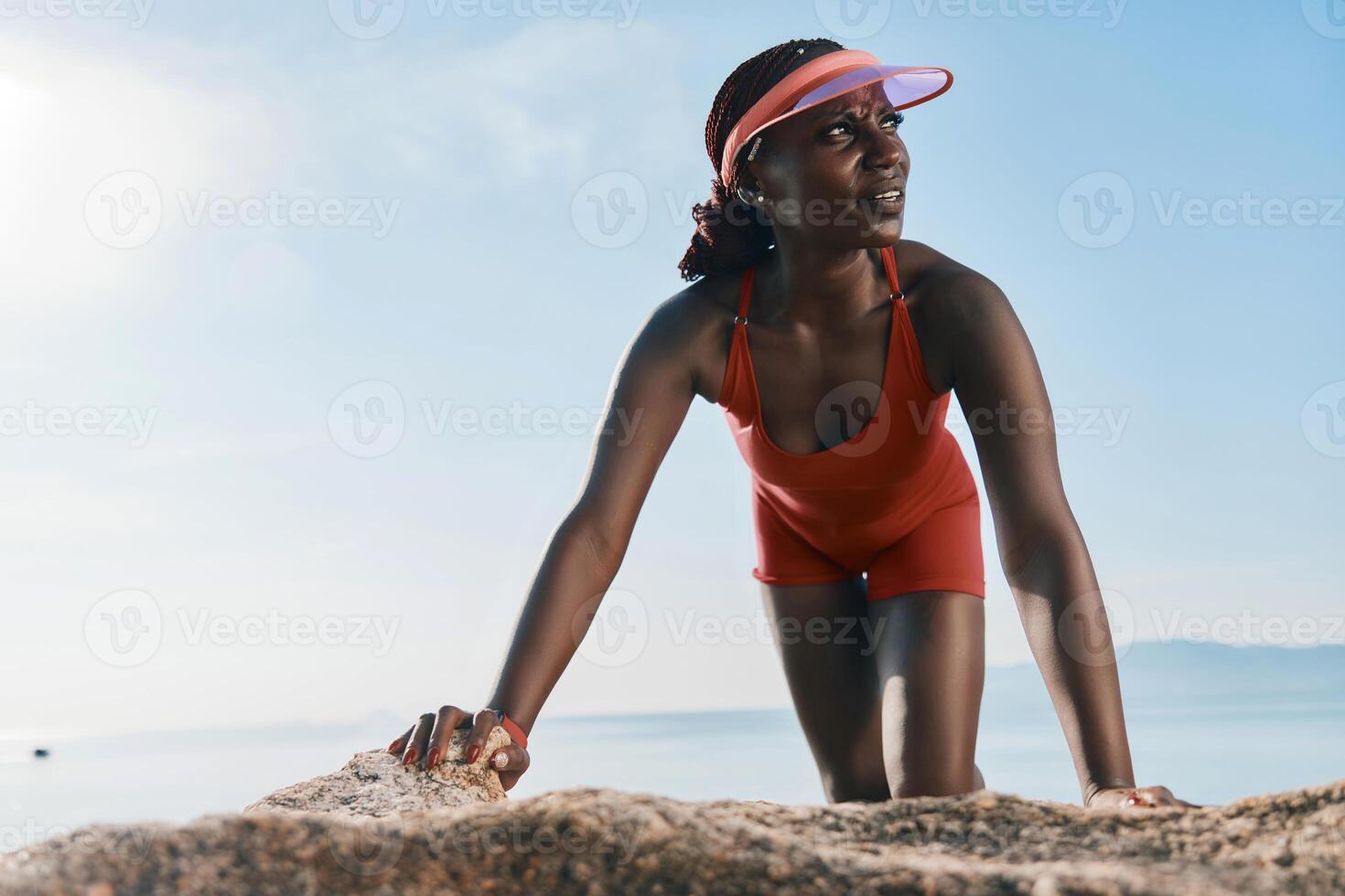 A fit woman in a vibrant red swimsuit stretches on rocky beach, showcasing determination and photo