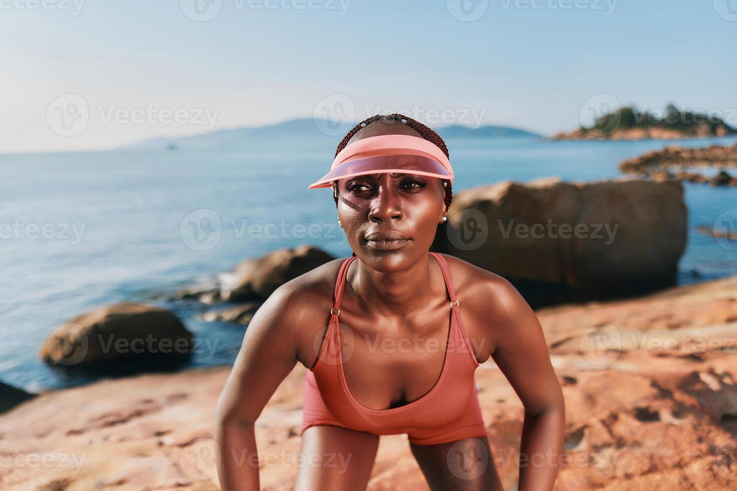 A focused woman in sportswear crouches on a rocky beach, with a serene ocean backdrop and soft photo