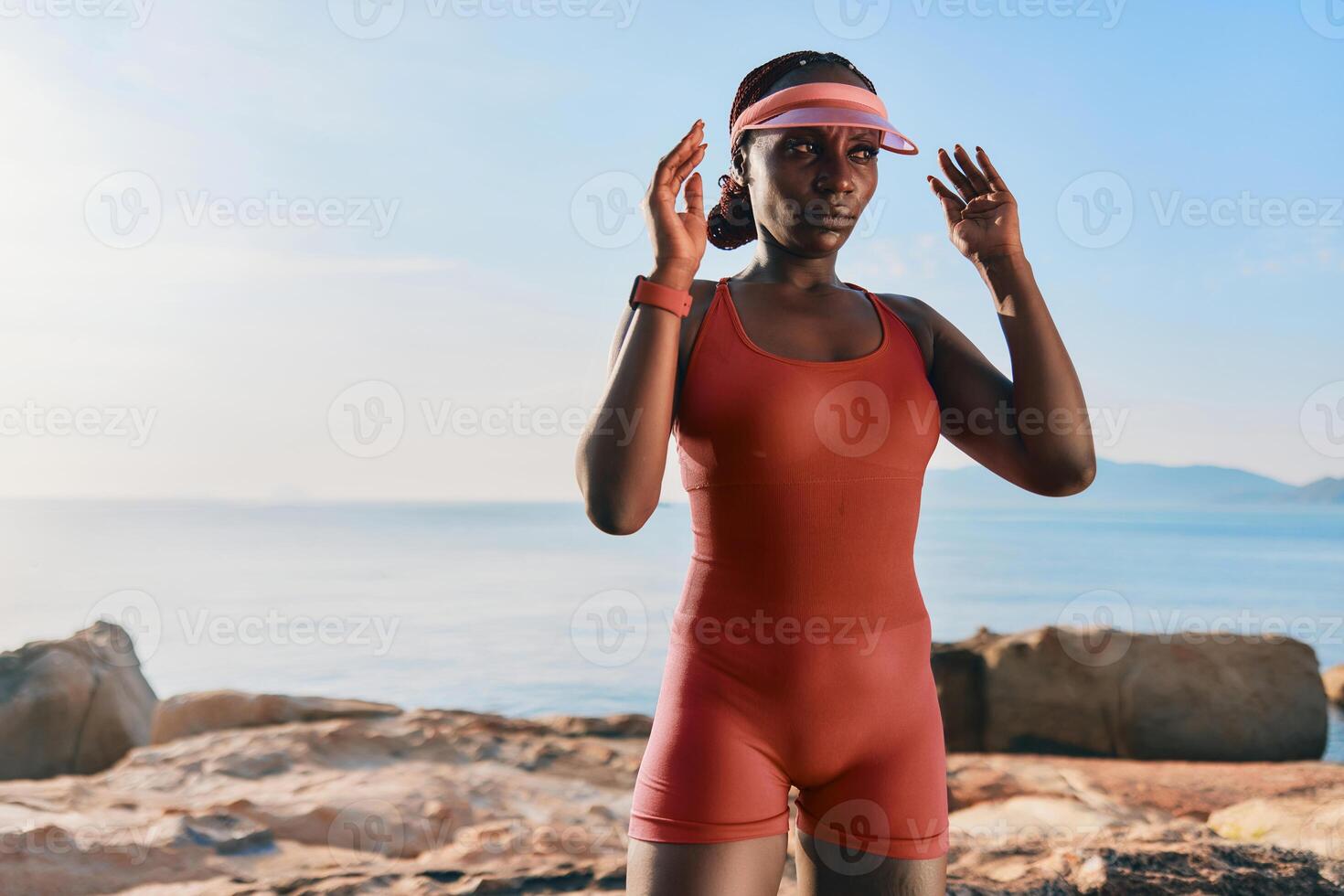 A fit woman in a red swimsuit poses confidently by the ocean, wearing a visor and enjoying the sunny photo