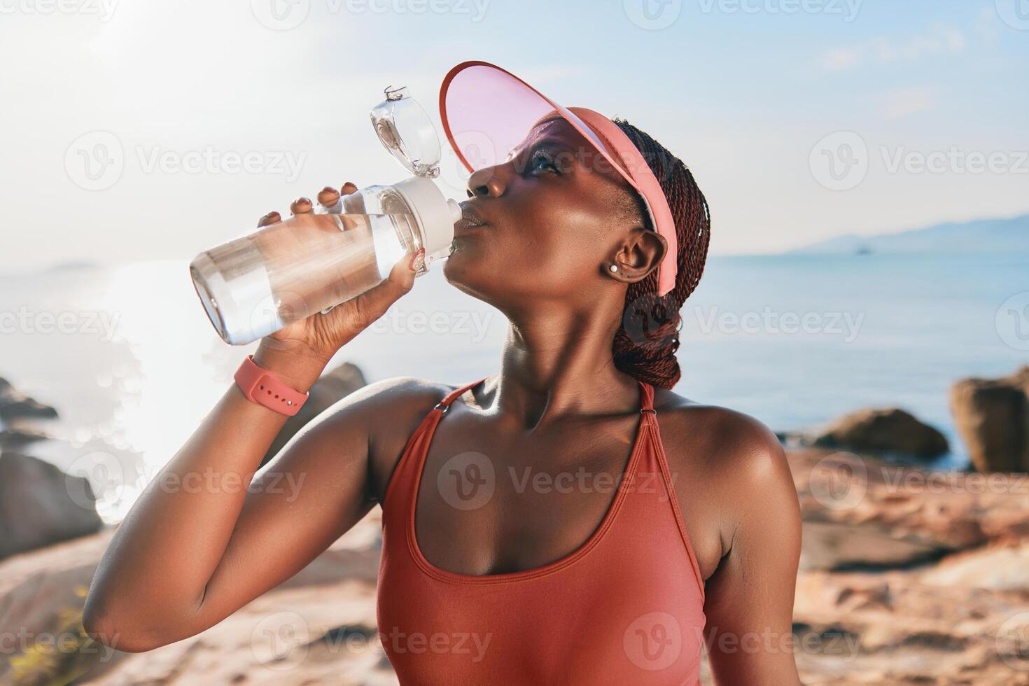 Athletic woman refreshing herself with water by the beach, wearing a cap and tank top, enjoying a photo