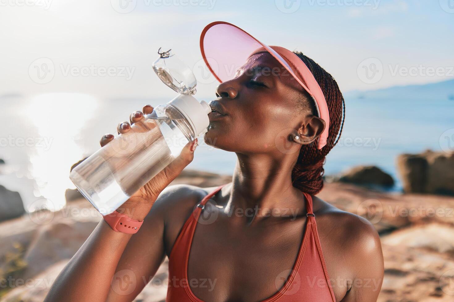 A fit woman in a cap enjoying water from a bottle, with a serene ocean background and a warm, sunny photo